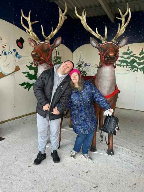 Two holidaymakers posing with reindeer figures in front of a snowy winter mural